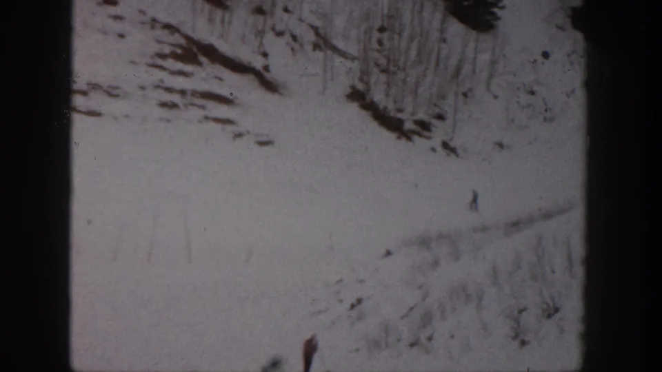 Distant 1961 Aspen ski scene with scattered figures on a pale snow slope beneath rocky, tree-dotted terrain.