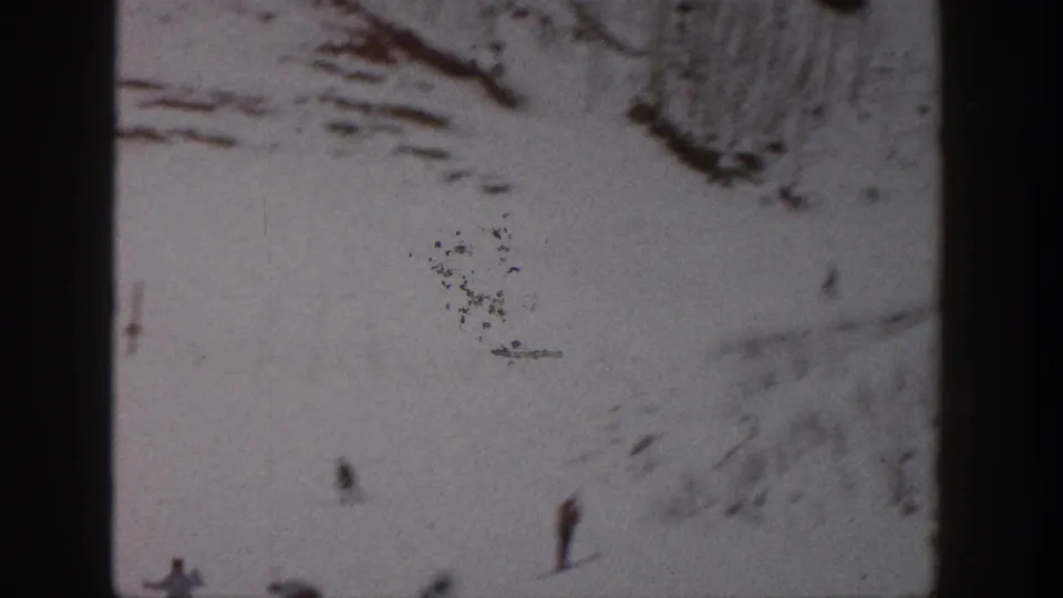 Grainy Aspen ski frame from 1961 with distant skiers, a wide snowfield, rocky background, and visible film wear.