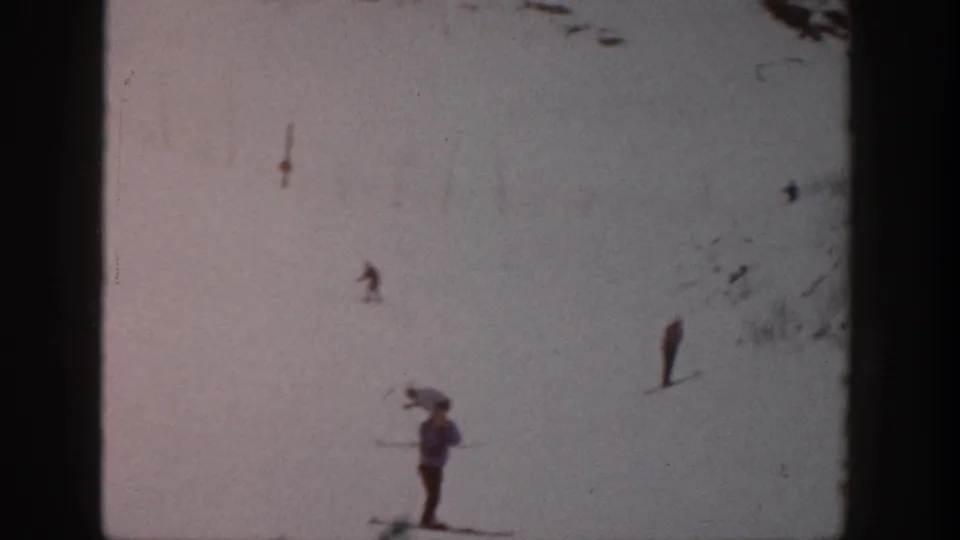 1961 Aspen home-movie still showing a closer skier near the lower frame and other small figures on the snowy hillside.