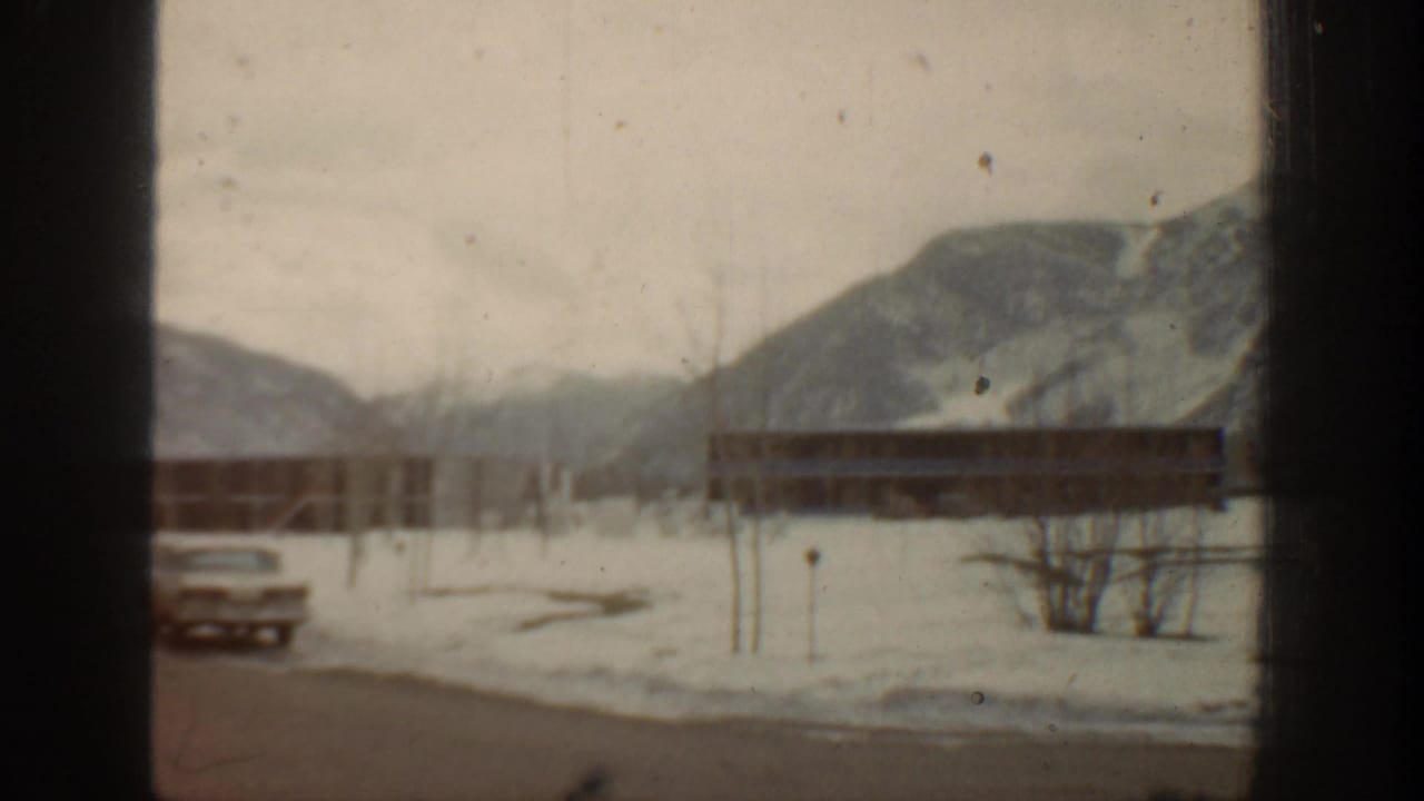A parked mid-century car sits near low buildings in snowy Aspen, with mountains in the background.