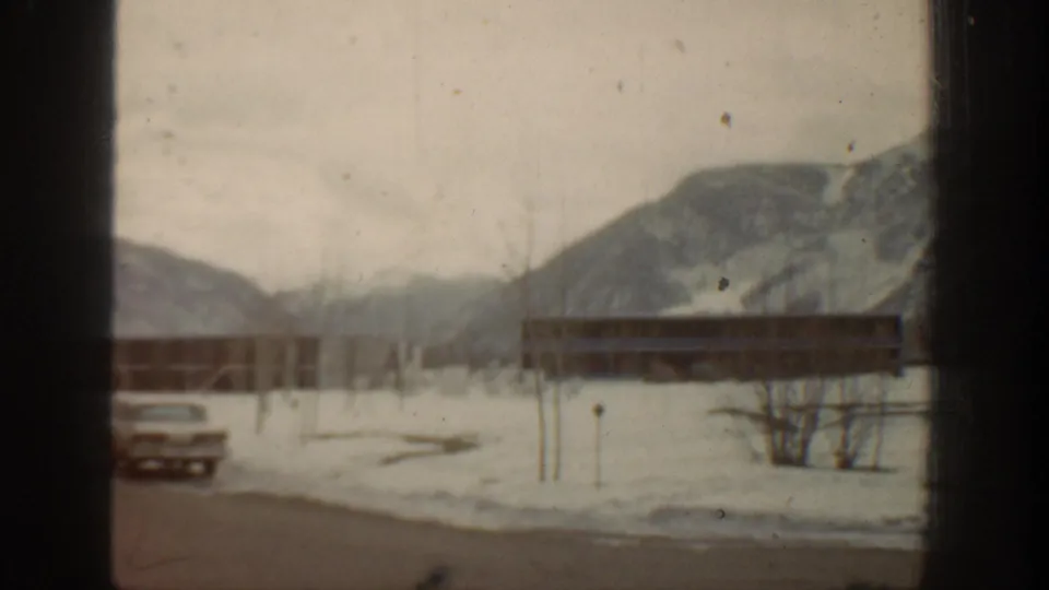 A parked mid-century car sits near low buildings in snowy Aspen, with mountains in the background.