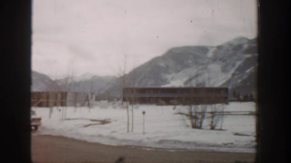 A soft 8mm view of snowy Aspen grounds, a low building, bare trees, and mountain ridges in 1961.