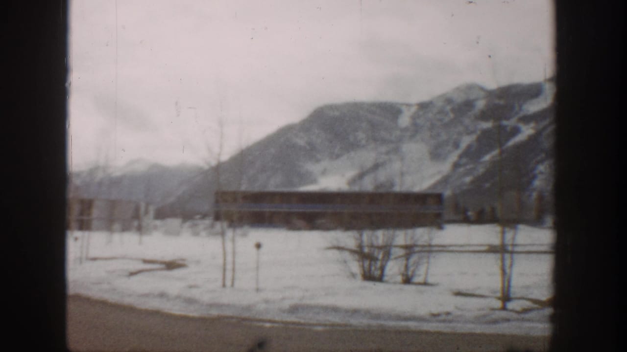 1961 Aspen home-movie frame showing a snowy open area, long low building, bare trees, and mountain backdrop through a window frame.