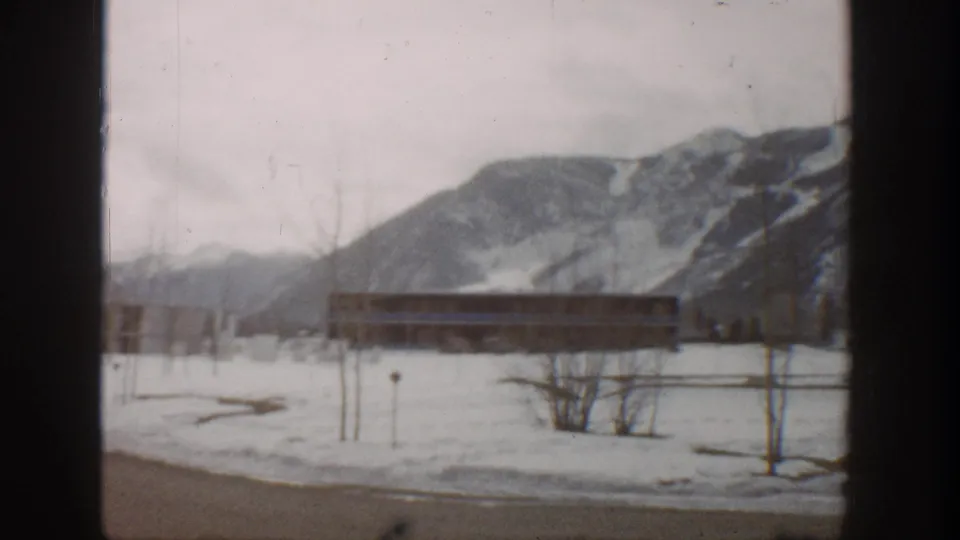 1961 Aspen home-movie frame showing a snowy open area, long low building, bare trees, and mountain backdrop through a window frame.