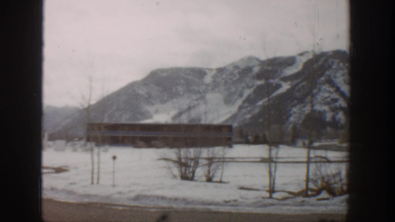 Snow-covered foreground, low rectangular building, bare trees, and mountain ridge behind window bars.