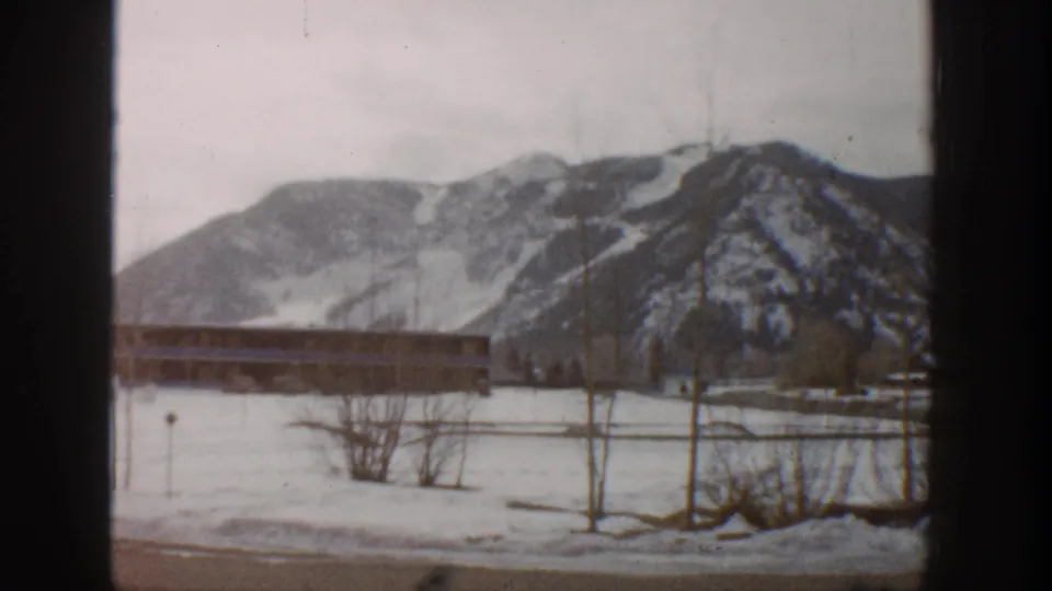 1961 Aspen 8mm frame with snow, a long low structure, bare winter trees, and rugged mountain slopes in the background.