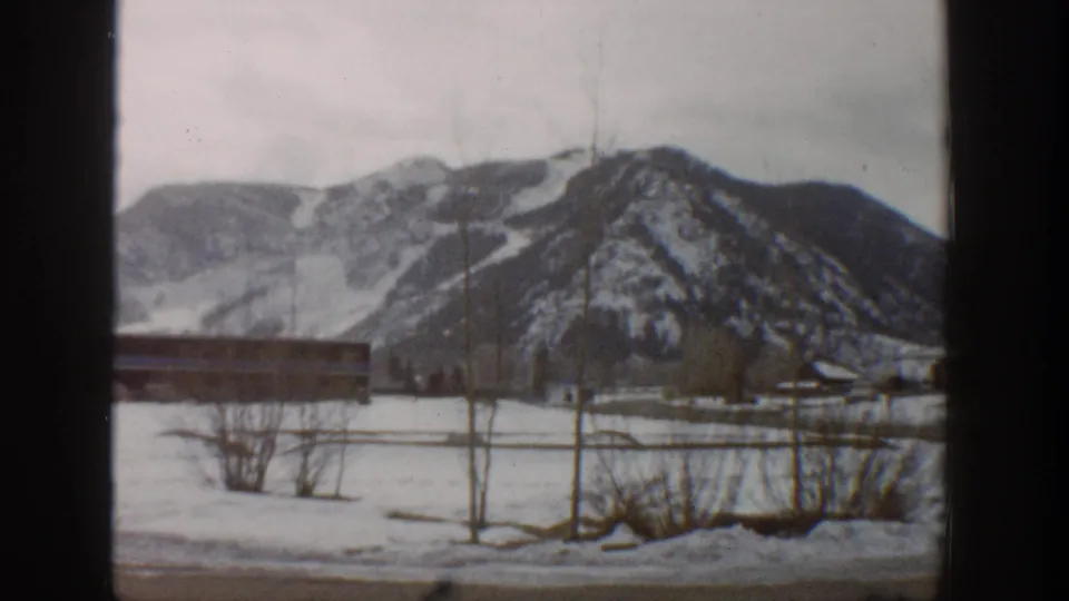 Final Aspen winter frame showing the same snowy field, low building, bare trees, and mountain backdrop through the window frame.