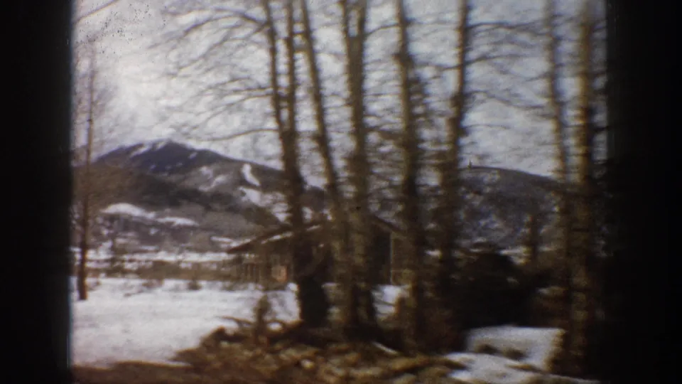 1961 Aspen, Colorado home-movie frame showing a snowy yard, leafless trees, mountain slopes, and a dark parked vehicle or structure.