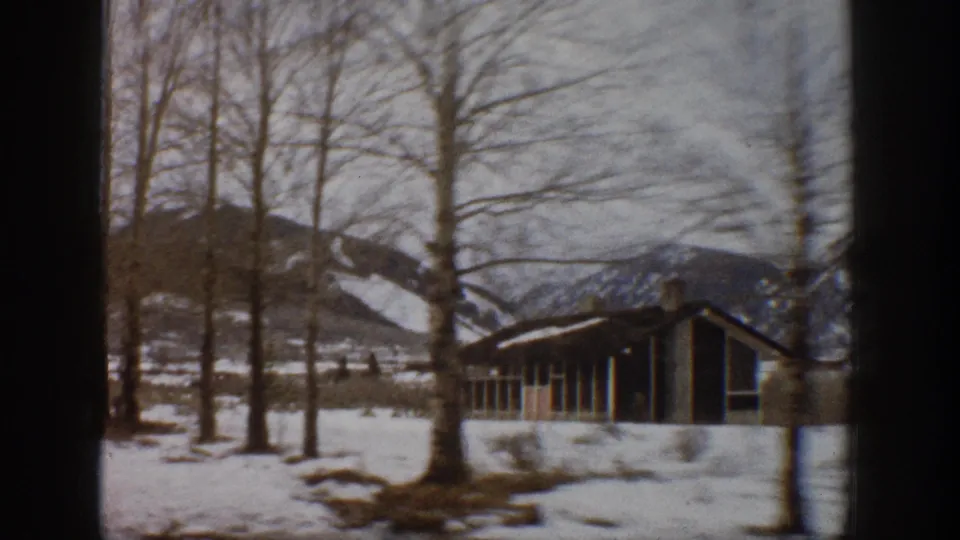 1961 Aspen, Colorado winter frame with a small cabin, bare trees, snow-covered ground, and mountains beyond.