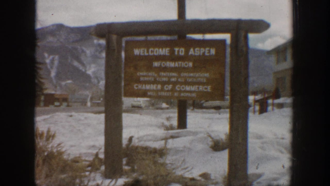 Wooden Welcome to Aspen information sign standing in snow with mountains behind it.