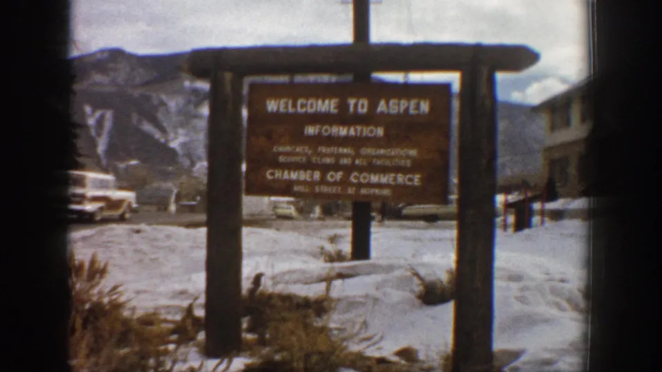 1961 Aspen, Colorado close frame of the Chamber of Commerce welcome sign with snowy roadside ground and alpine backdrop.