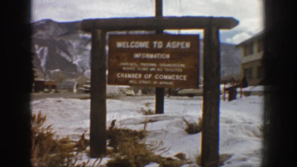1961 Aspen, Colorado home-movie frame centering the welcome sign, with snow, parked cars, buildings, and mountains behind it.