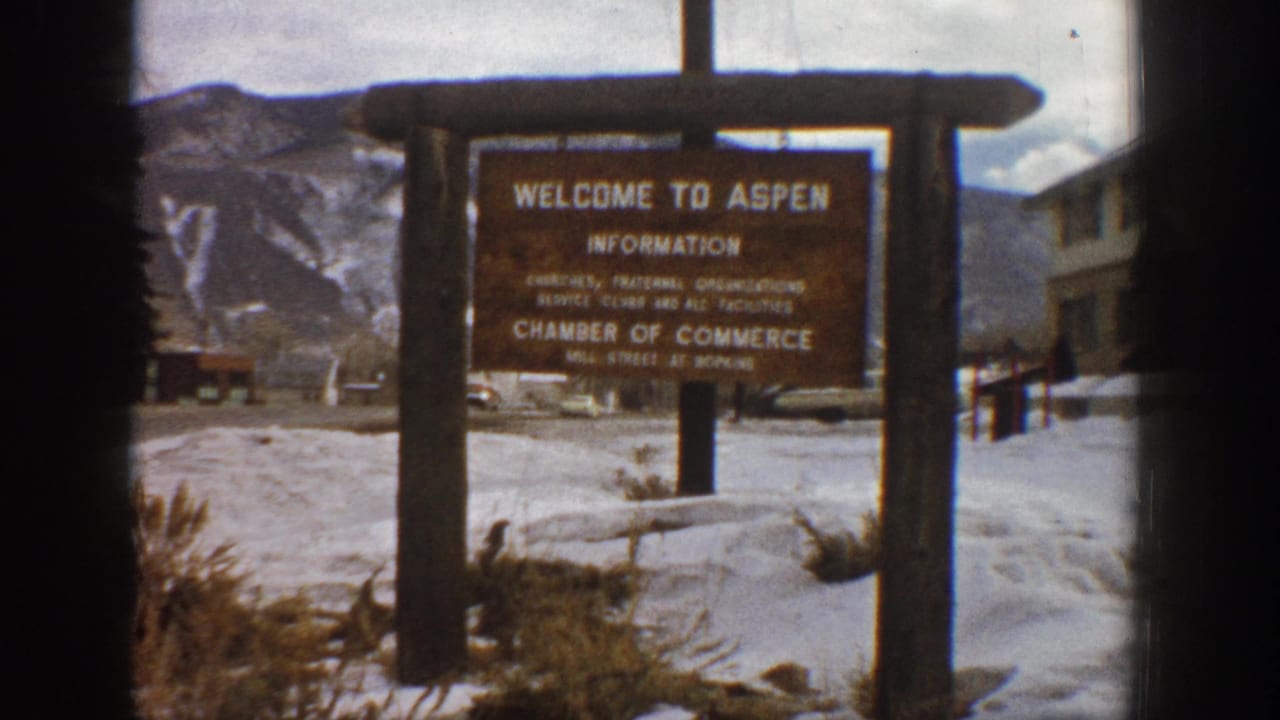 1961 Aspen, Colorado home-movie frame showing a wooden Welcome to Aspen information sign in a snowy roadside setting with mountain slopes in the background.