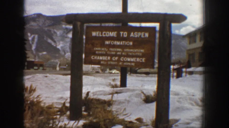 A slightly closer 1961 Aspen frame holds on the wooden welcome sign, snowy ground, mountain slopes, and nearby roadside buildings or parked cars.