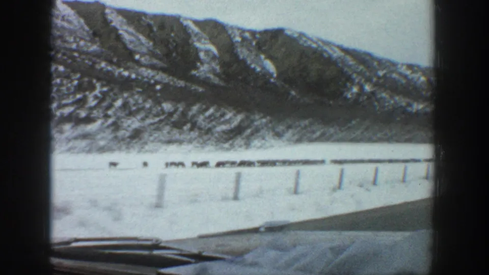 Moving-vehicle view in Aspen, Colorado showing snow-dusted mountain slopes, a snowy roadside, fence posts, and part of the car windshield or dashboard.