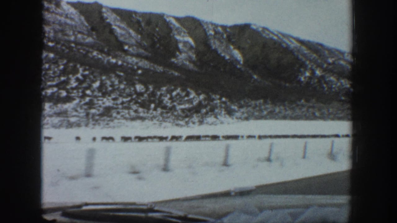 1961 Aspen, Colorado 8mm frame showing a snowy road from inside a car, with mountains, fence posts, and livestock across the field.