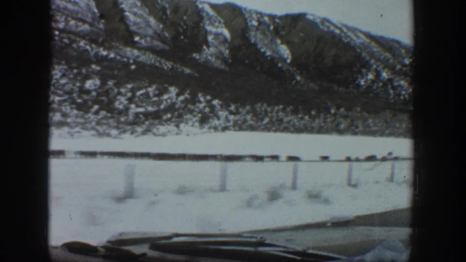 1961 Aspen, Colorado 8mm still showing a moving car view of a snow-covered field, fence posts, livestock, and mountain slopes.