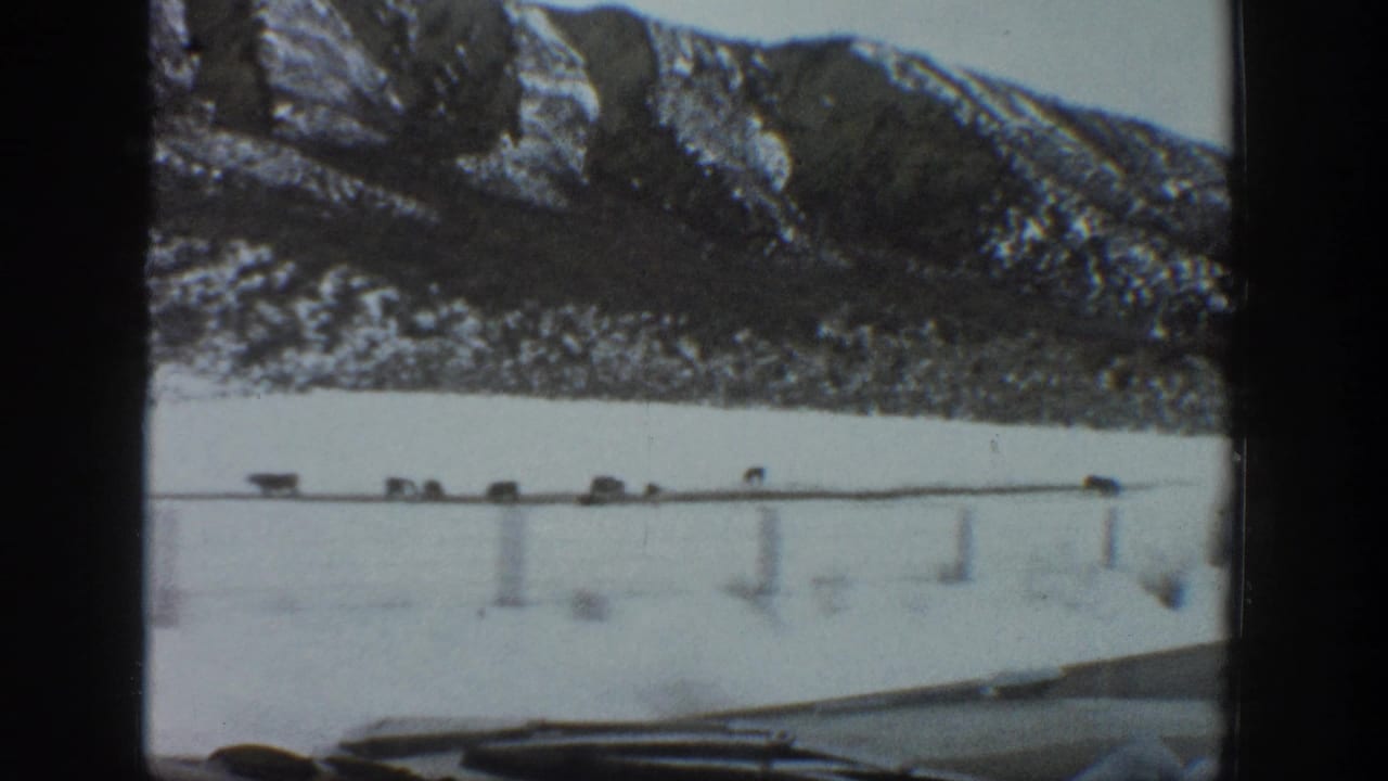 1961 Aspen-area 8mm frame showing a snow-covered road, small vehicles, dashboard edge, and steep winter mountains beyond.
