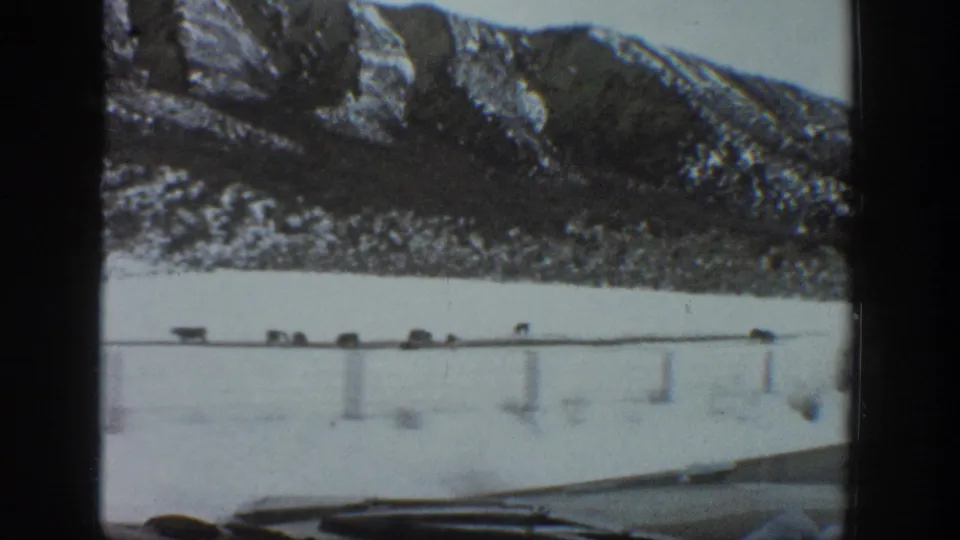 1961 Aspen-area 8mm frame showing a snow-covered road, small vehicles, dashboard edge, and steep winter mountains beyond.