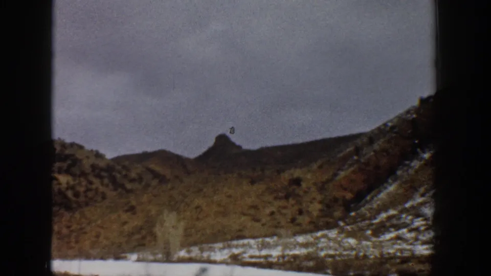 Aspen-area winter landscape from 1961 8mm footage, with snow patches, brown hillsides, and a pointed ridge under gray sky.