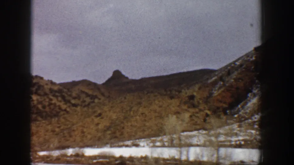 Aspen-area 1961 8mm road-trip view showing snow along the roadside, scrub-covered hills, and a distant pointed mountain form.