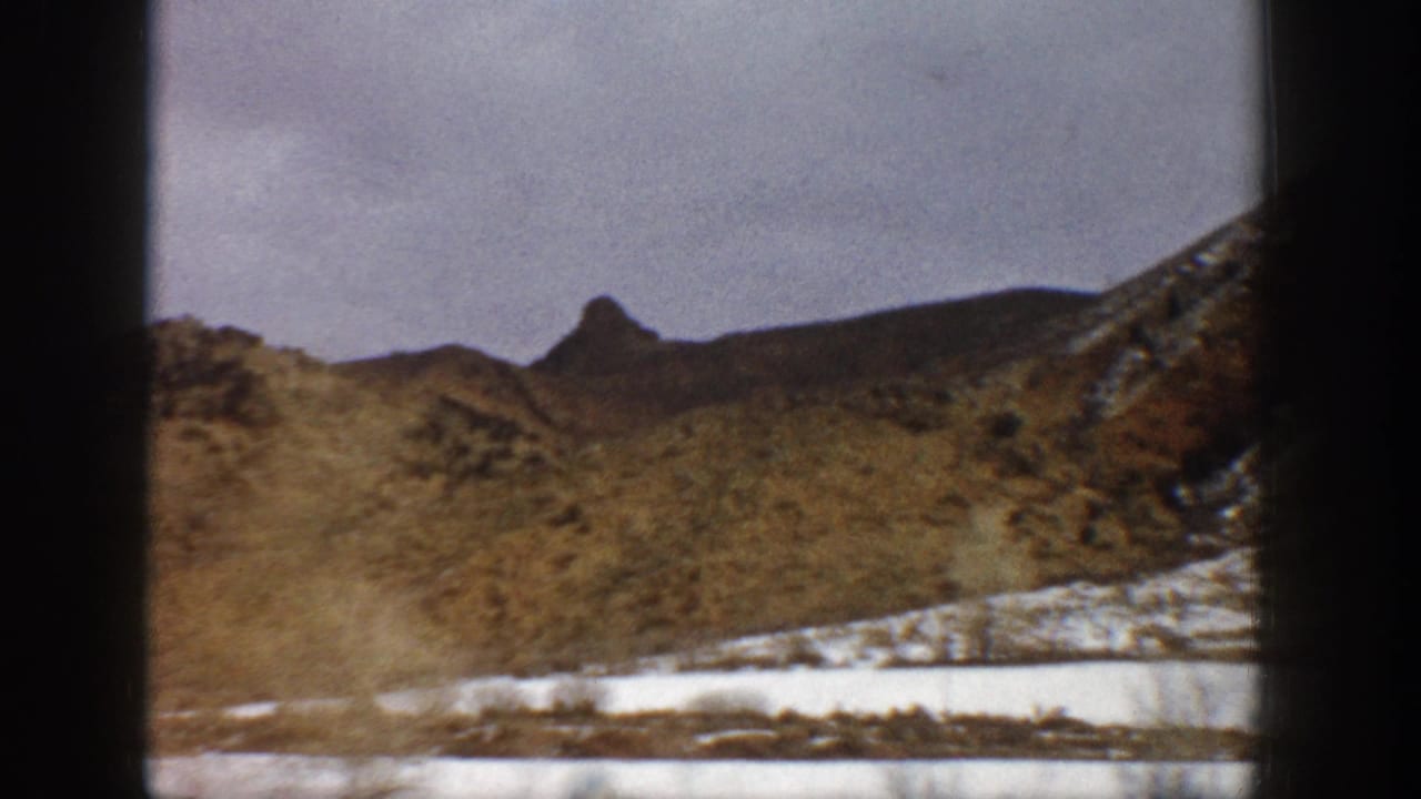 1961 Aspen-area home-movie view of snow patches across a field and brown hills, framed by dark edges from a moving vehicle.