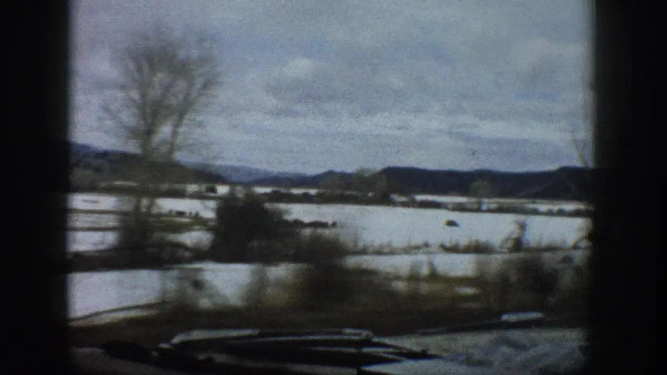 A moving-vehicle view near Aspen in 1961 shows snowy open ground, bare roadside trees, low hills, and part of the car hood.