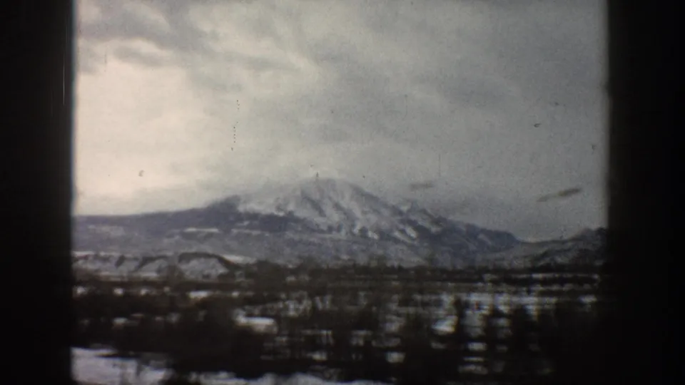 1961 Aspen-area frame with a snow-streaked mountain ridge, dark winter trees, open valley ground, and cloudy sky in soft 8mm color.