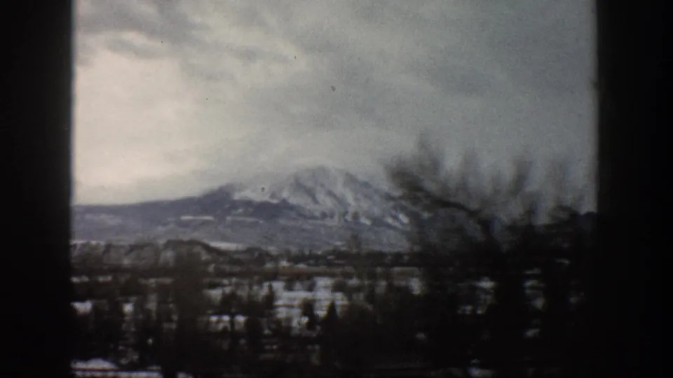 1961 Aspen, Colorado home-movie view of a snowy mountain ridge, bare branches, valley trees, and gray sky from inside a moving car.