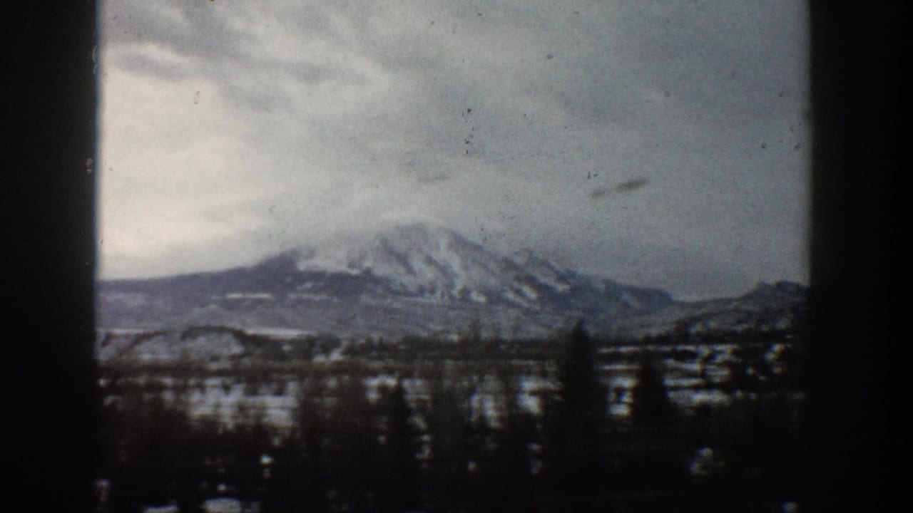 Vintage 8mm view of Aspen-area snowy mountains, with a dark tree line in the foreground and muted winter clouds overhead.