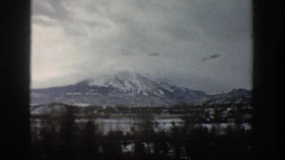 1961 Aspen, Colorado home-movie frame with snow-striped mountains, foreground trees, cloudy sky, and visible 8mm gate edges.