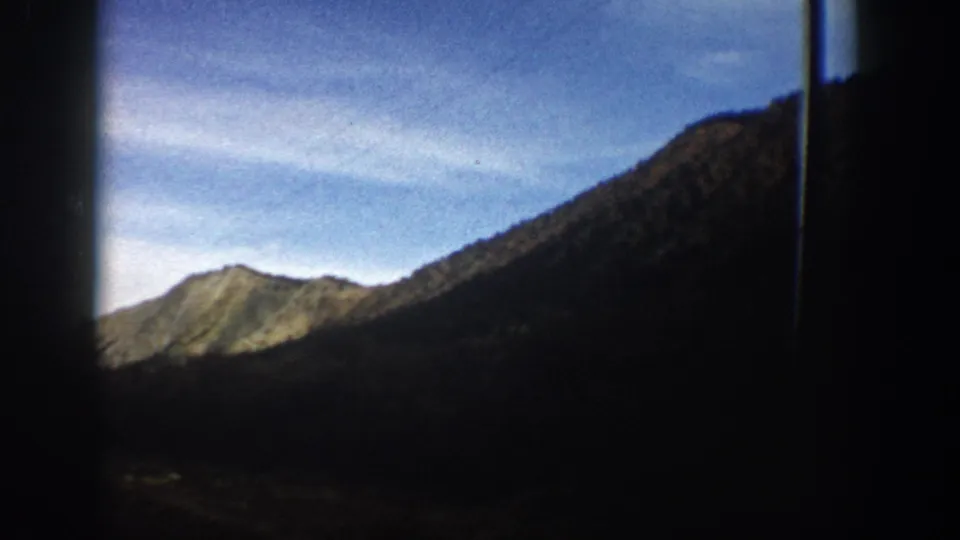1961 Colorado home-movie frame showing a window-framed mountain view, with a sunlit peak, high ridge, and dark valley foreground.