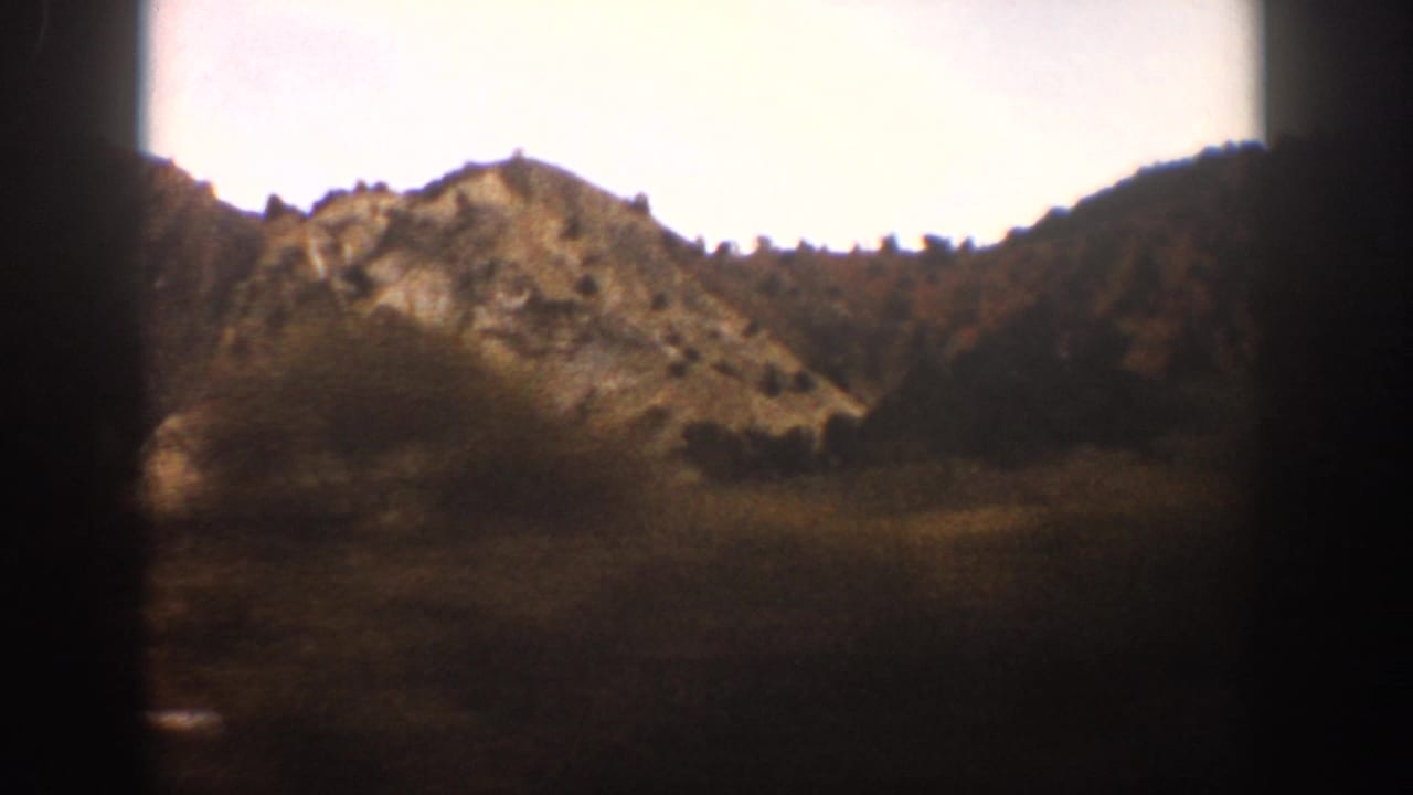 A warm-toned 8mm view from a train near Aspen shows a rocky ridge, scattered trees, open land, and dark window framing.