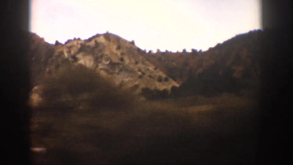 A warm-toned 8mm view from a train near Aspen shows a rocky ridge, scattered trees, open land, and dark window framing.