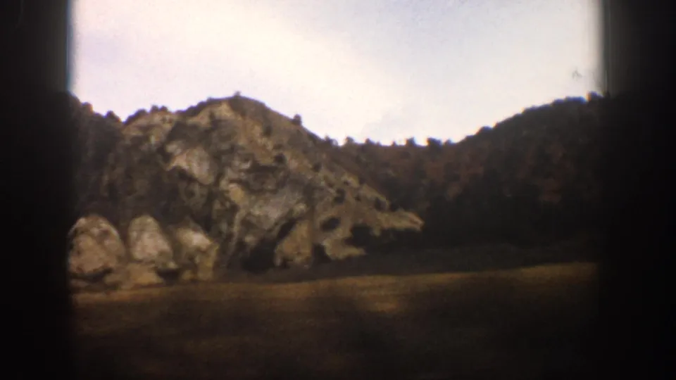 The 1961 home-movie frame shows rock outcrops, a sloped tree line, field, and dark window edges during a train ride near Aspen.