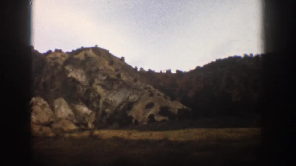 A train-window view near Aspen captures a rocky hillside, scattered trees, open field, and pale sky in soft 1960s 8mm color.