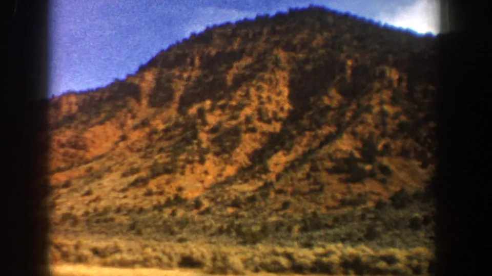 1961 Aspen-area driving footage with a closer view of a rocky, brush-covered hillside through the vehicle window.