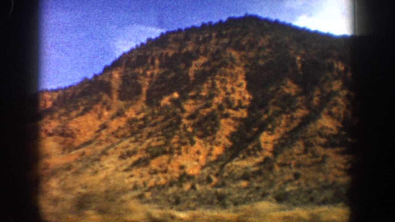 1961 Aspen-area 8mm frame showing a sunlit rocky mountainside, sparse vegetation, and a dark treeline beneath blue sky.