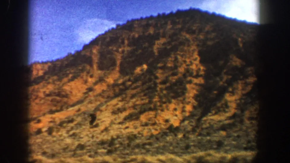 1961 Colorado 8mm frame of a rugged orange-brown hillside with scattered brush and a dark wooded ridge line.