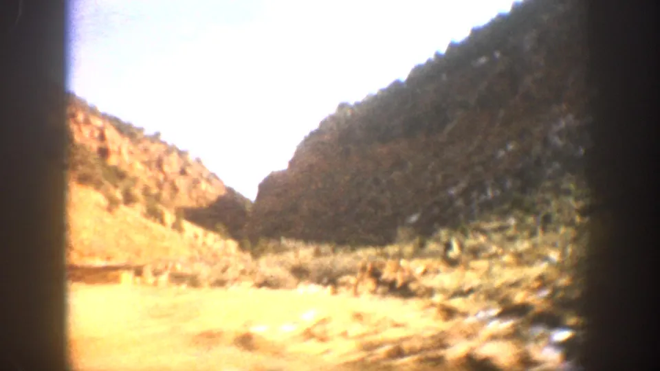 1961 Aspen-area 8mm frame from a moving vehicle, showing a blurred canyon slope, dry roadside ground, and bright sky.