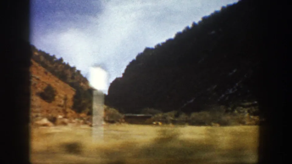 1961 Colorado 8mm frame showing a wider canyon view with dark slopes, dry foreground, cloudy sky, and a distant pale structure-like form.