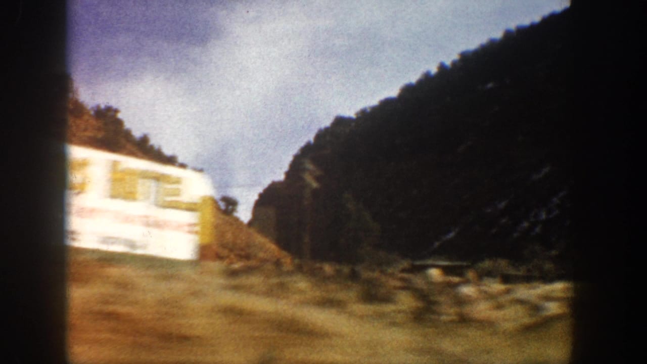 1961 Aspen-area 8mm frame from a moving vehicle, showing a pale roadside object, sloped canyon terrain, trees, and dark film borders.