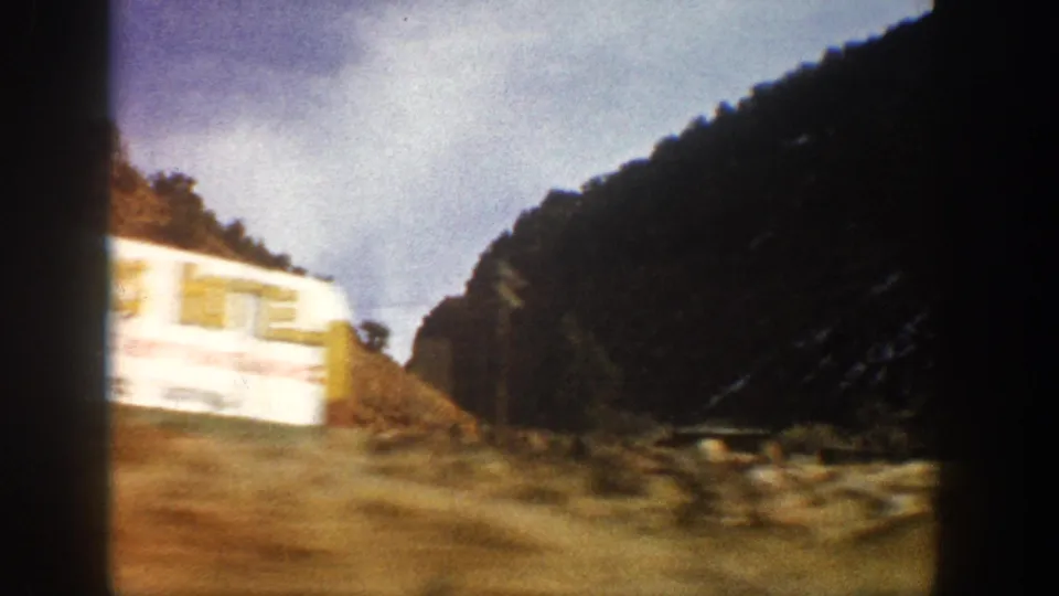 1961 Aspen-area 8mm frame from a moving vehicle, showing a pale roadside object, sloped canyon terrain, trees, and dark film borders.