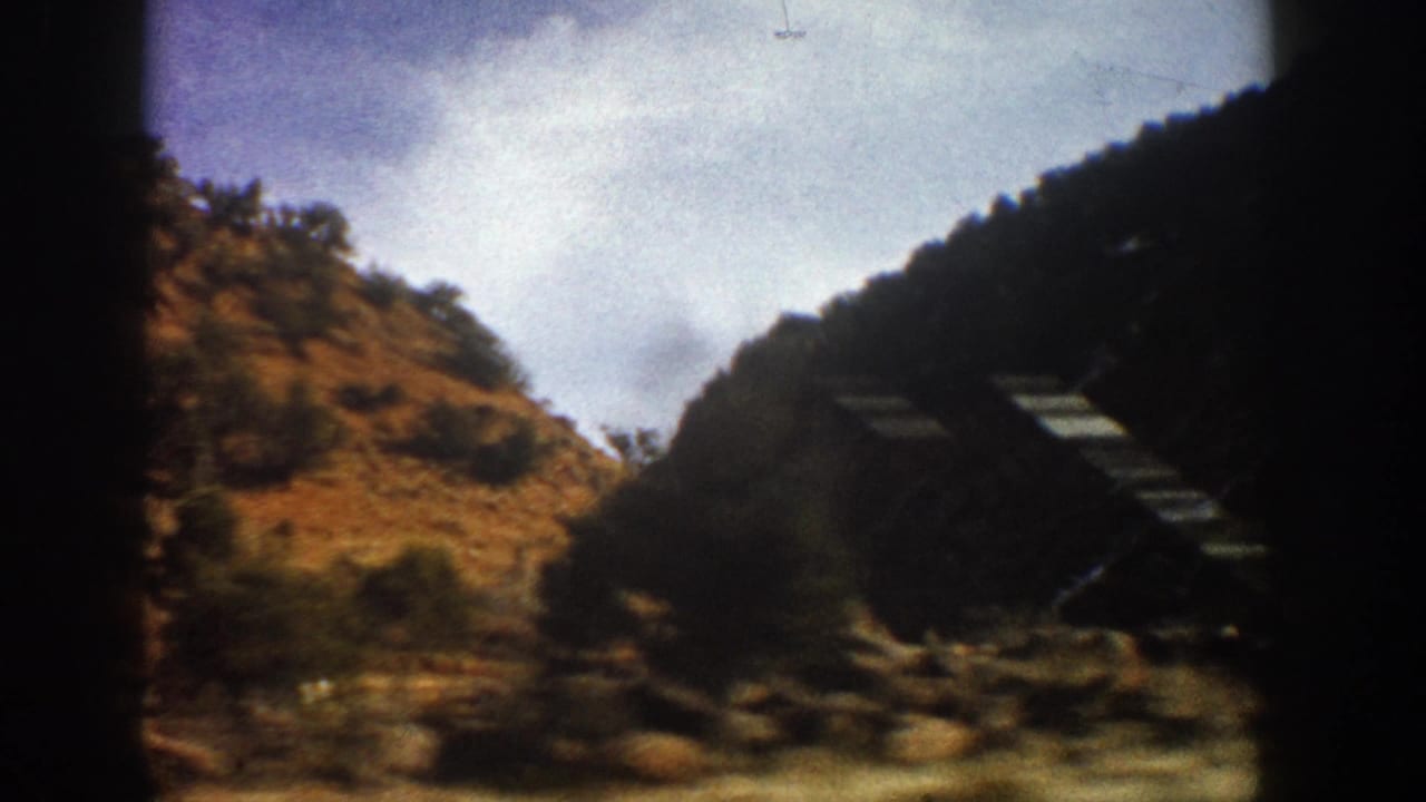Aspen-area mountain drive frame showing rocky terrain, tree cover, bright sky, and a dark diagonal roadside form blurred by motion.