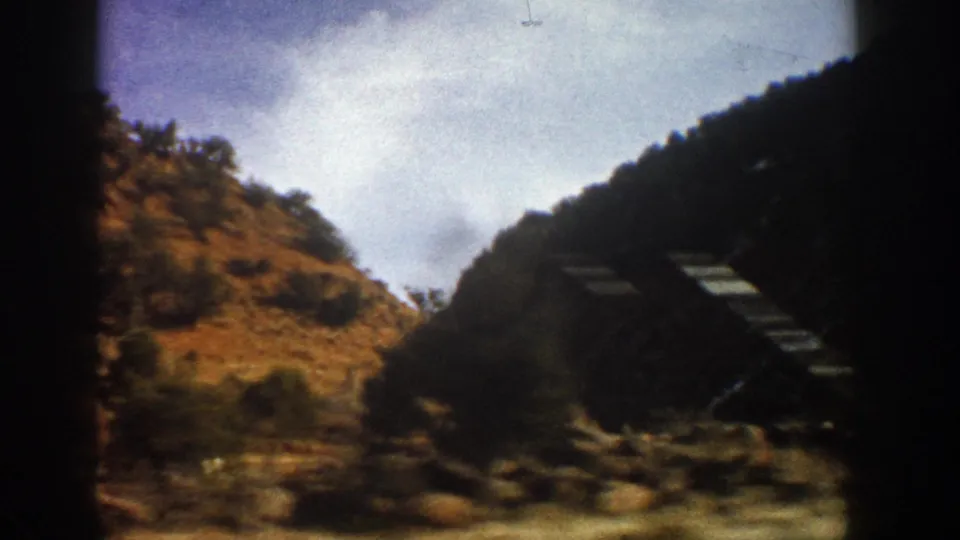 Aspen-area mountain drive frame showing rocky terrain, tree cover, bright sky, and a dark diagonal roadside form blurred by motion.