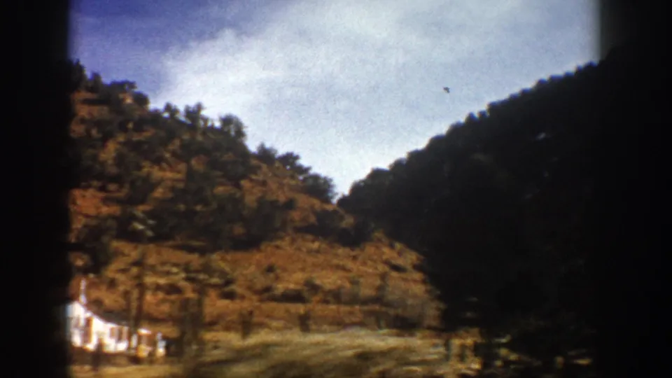 1961 Colorado home-movie frame from a vehicle, with canyon slopes, roadside brush, trees, sky, and a pale object near the lower left edge.