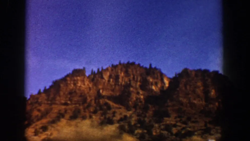 Aspen-area 8mm frame showing a sunlit rocky ridge, sparse trees, blue sky, and rounded dark borders from the film gate.