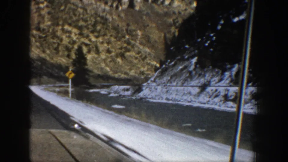 Aspen, Colorado 8mm frame from 1961 showing a mountain highway, snowbanks, rocky slopes, evergreen trees, and a yellow road sign.