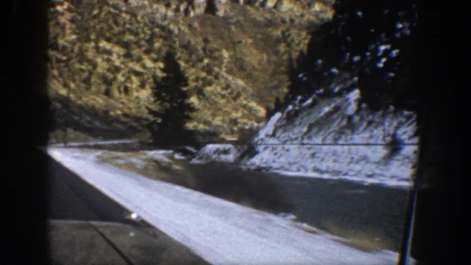1961 Aspen, Colorado home-movie frame showing snow along a mountain road, rocky hillside, dark evergreen trees, and the edge of a car window.
