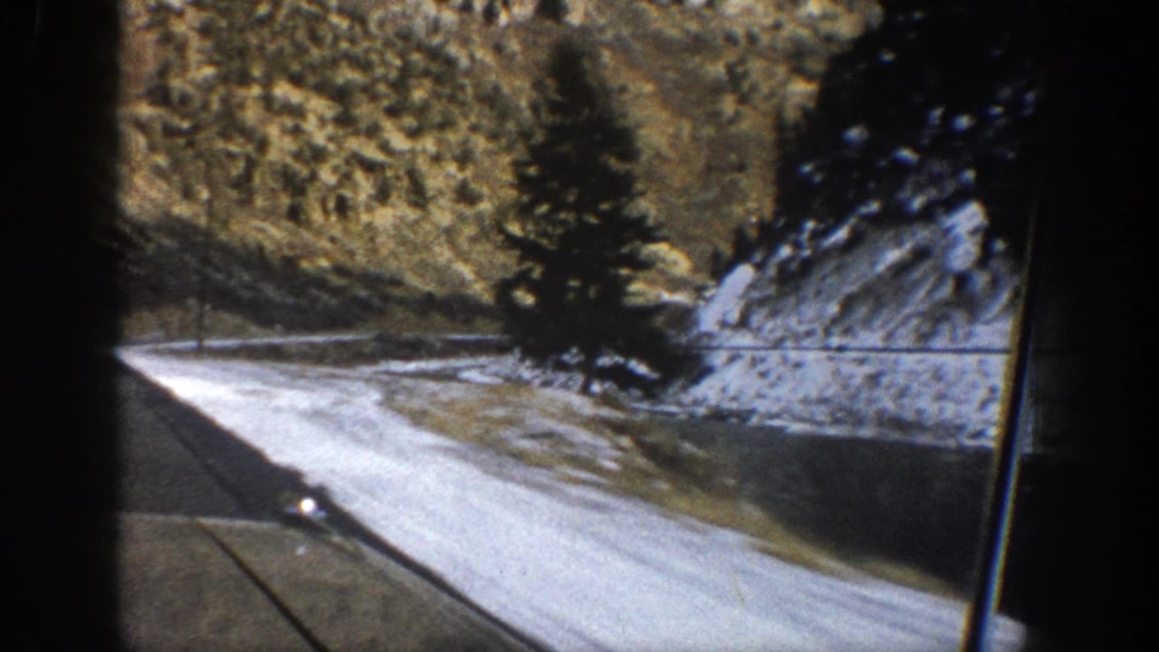 1961 Aspen-area mountain road filmed from inside a moving car, with snow along the roadside and a windshield wiper in view.